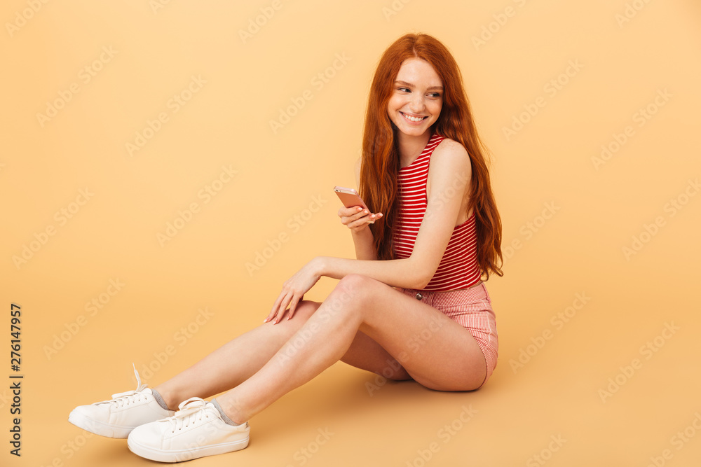 Smiling young beautiful redhead woman posing isolated over yellow background using mobile phone Smiling young beautiful redhead woman posing isolated over yellow background using mobile phone