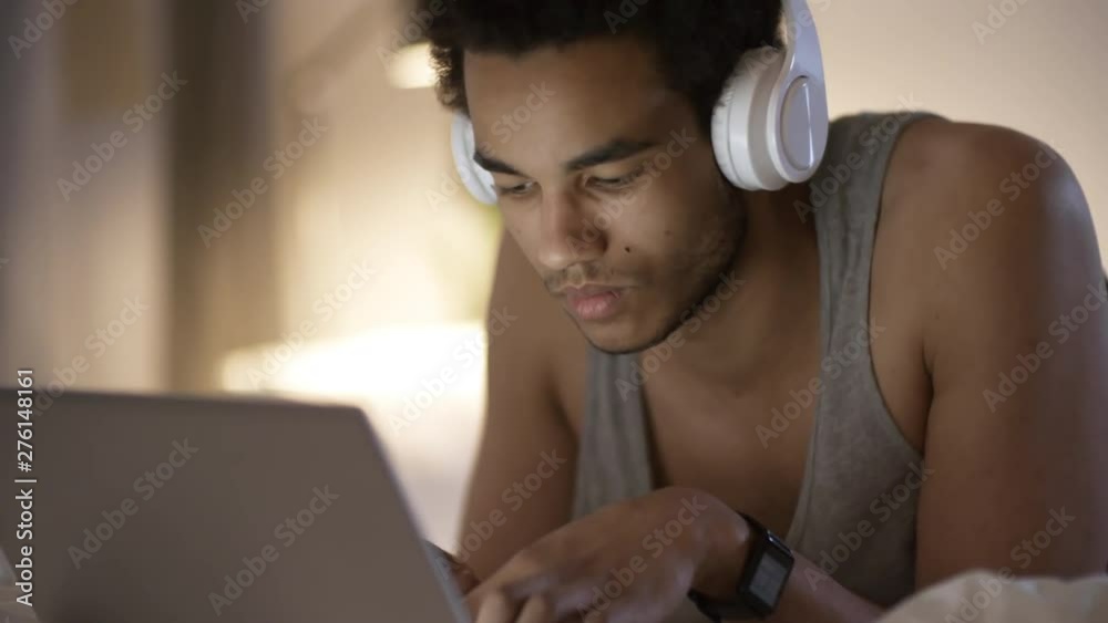 Close up shot of young black man with short curly hair lying in bed late at night, using laptop computer and listening to music with wireless headphones