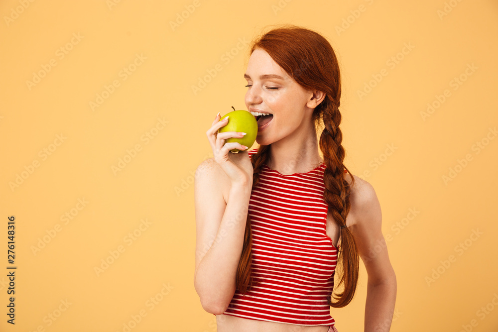 Happy young beautiful redhead woman posing isolated over yellow background eat apple. Happy young beautiful redhead woman posing isolated over yellow background eat apple.