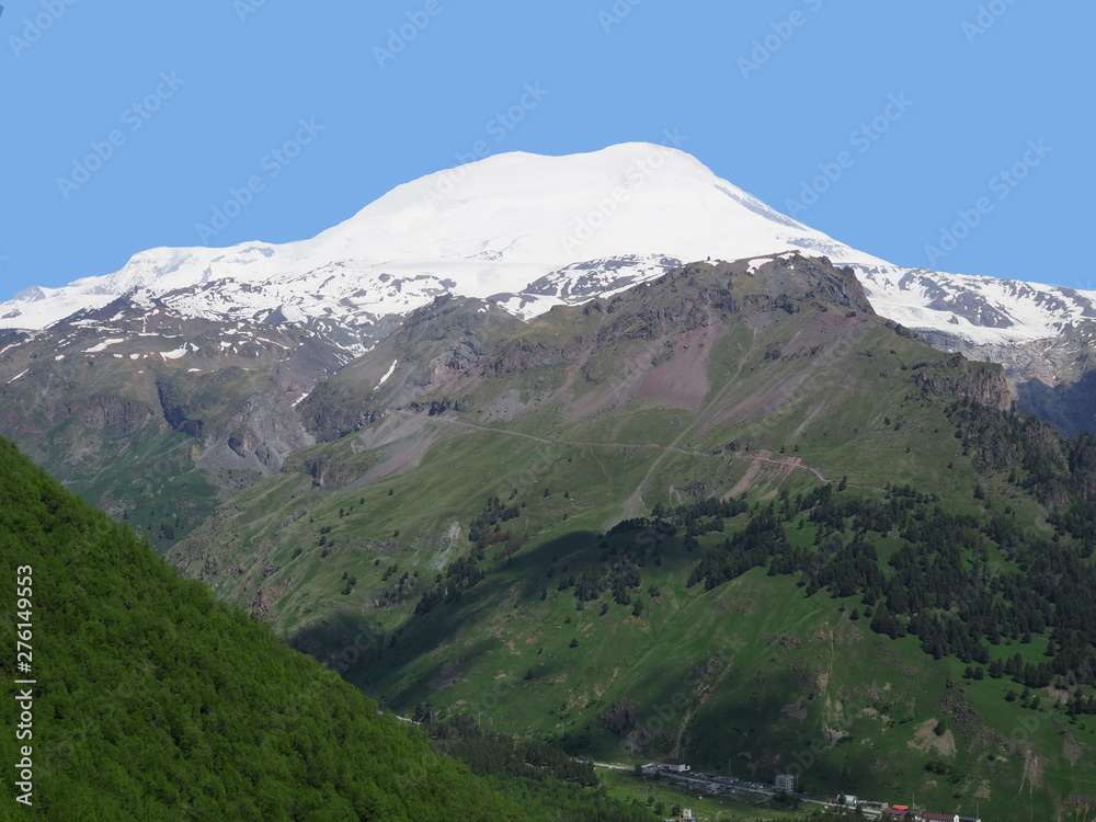 Great nature mountain range. Amazing perspective of caucasian snow mountain or volcano Elbrus with green fields, blue sky background. Elbrus landscape view - the highest peak of Russia and Europe