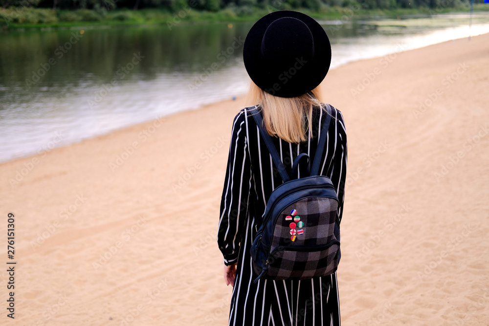 Fototapeta premium Blonde girl in black hat with backpack walking on the street. City traveling alone concept