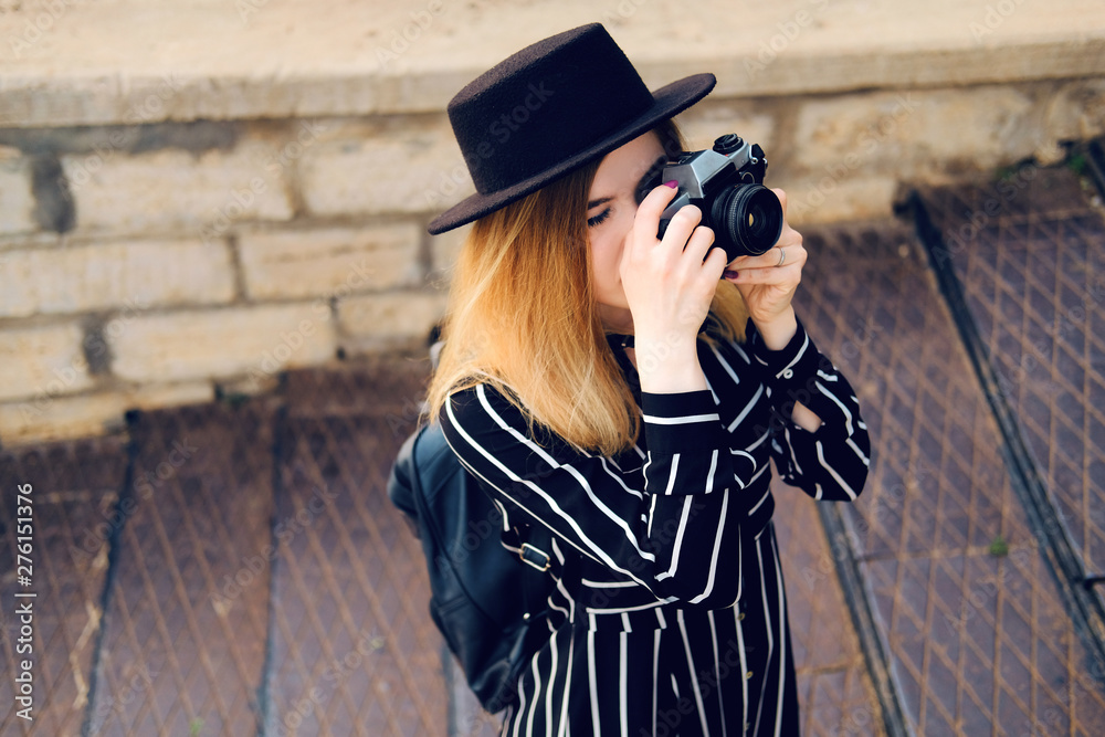 Blonde girl in black hat with backpack taking pictures with retro style camera. City traveling alone concept