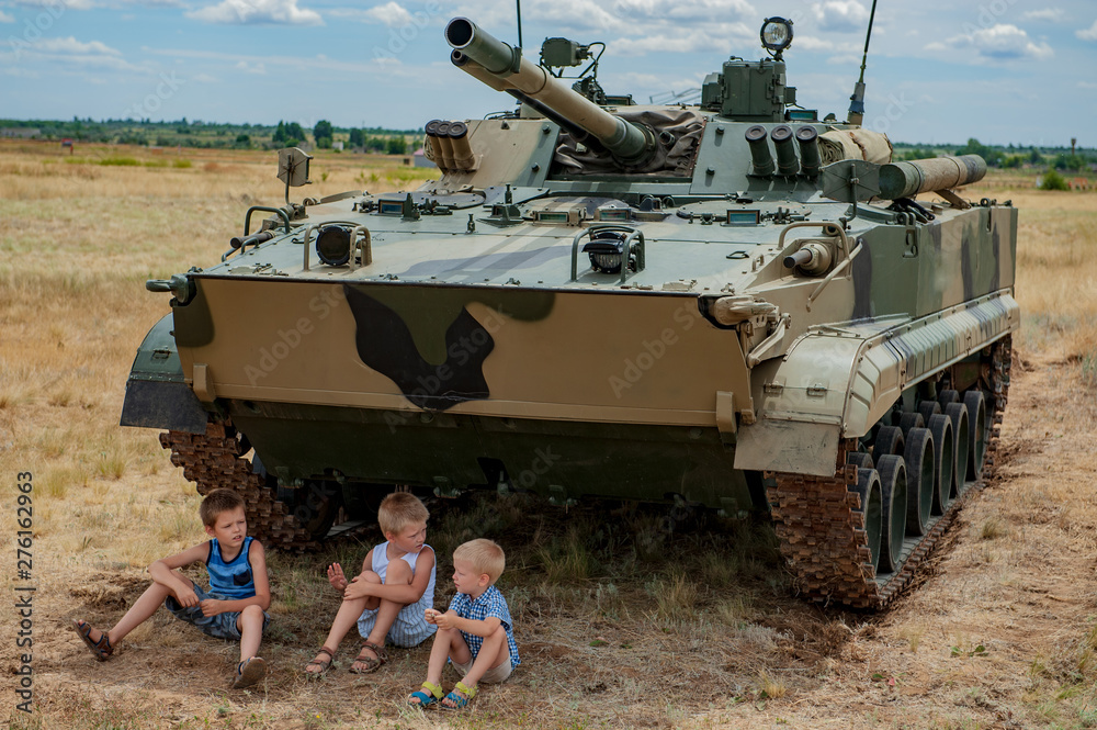 Three children in bright clothes are sitting in shade under modern battle tank. Children play until arrival of tankists. concept of contrasting innocent childhood and war