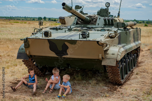 Three children in bright clothes are sitting in shade under modern battle tank. Children play until arrival of tankists. concept of contrasting innocent childhood and war