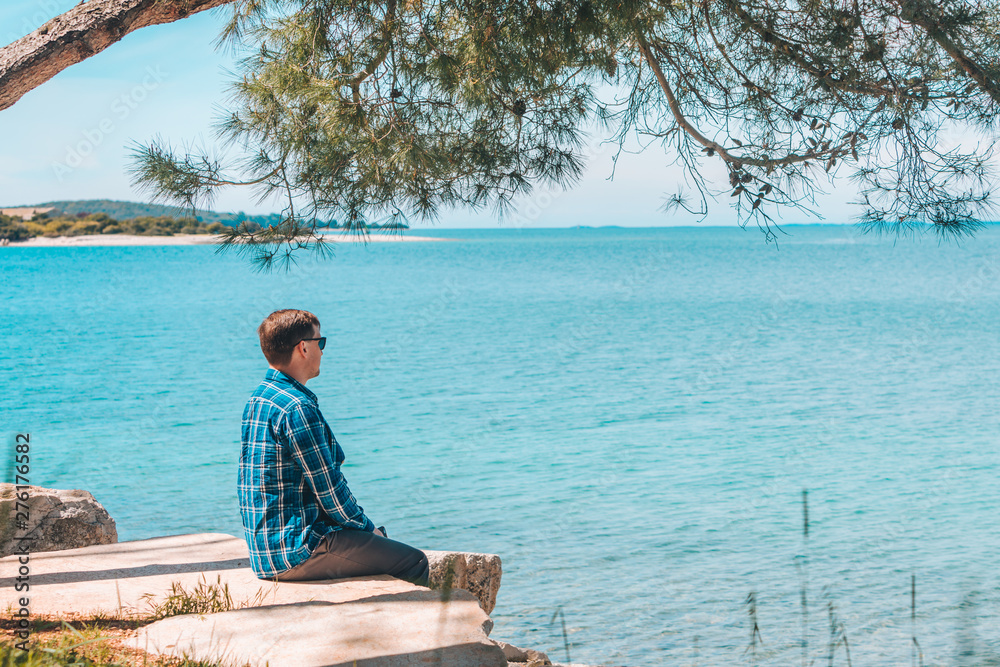 Fototapeta premium man sitting in the shadow of tree looking at blue azure water in sunny day