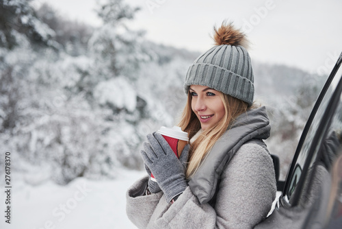 Warming up by fresh beverage. Pretty girl in warm clothes standing in the winter wood while leans on the car and holding cup of coffee