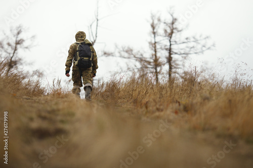 Traveler in camouflage clothes with a backpack rises up the hillside against the background of the autumn landscape. View from the back.
