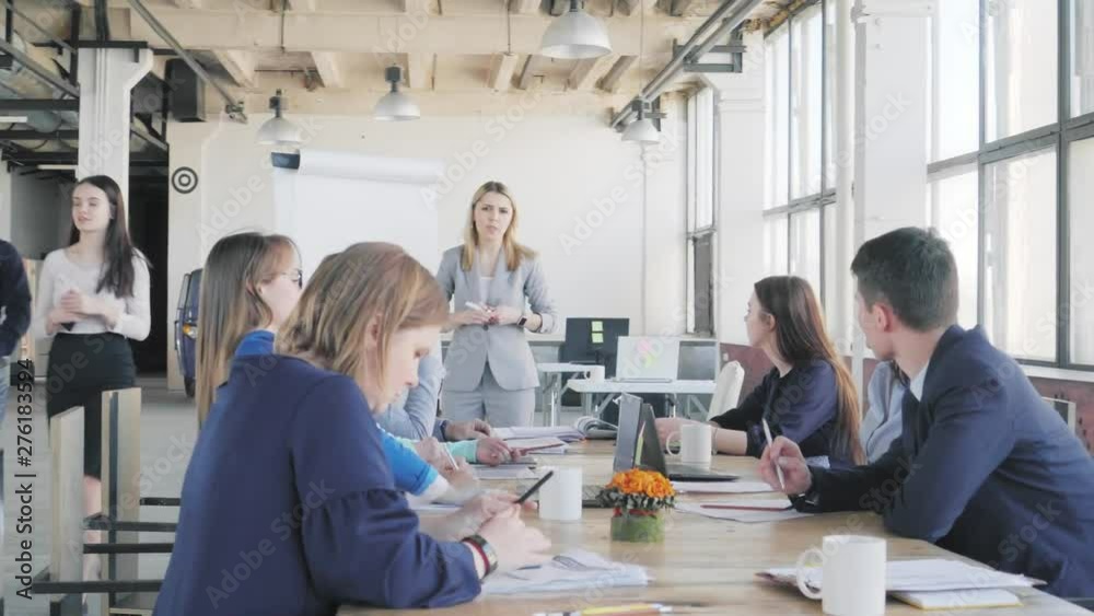 The team leader conducts a presentation near a large table and a flipchart and describes business processes. Employees sit at a table. Office life in the background. Coworking The team leader conducts a presentation near a large table and a flipchart and describes business processes. Employees sit at a table. Office life in the background. Coworking