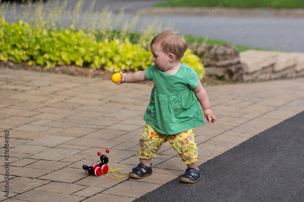 Side view of adorable fair toddler girl in summer clothes holding an egg-shaped object while standing in a driveway with toys Side view of adorable fair toddler girl in summer clothes holding an egg-shaped object while standing in a driveway with toys