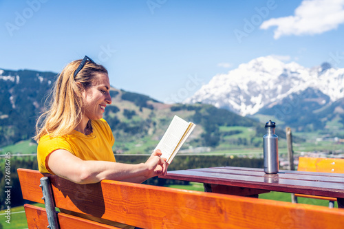Young beautiful and happy girl reading a book on a bench, mountain range and greenery in the background.