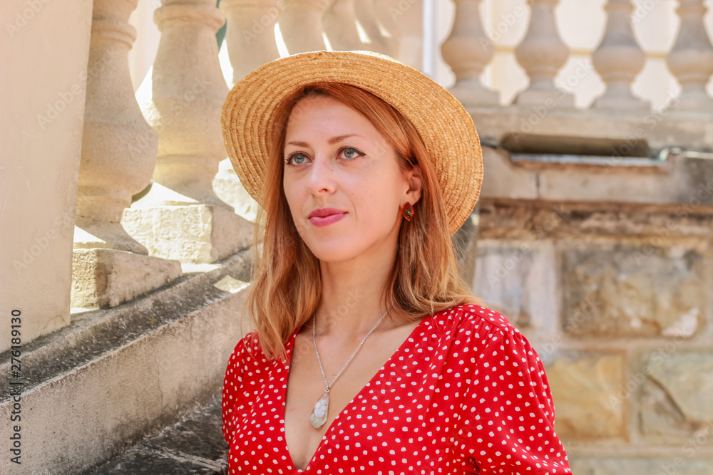 Photosession model with decorations of epoxy resin. Bijouterie. Red dress in peas. Straw hat. Stylish images. Fashion. Summer Trends 2019. Old architecture in the background.