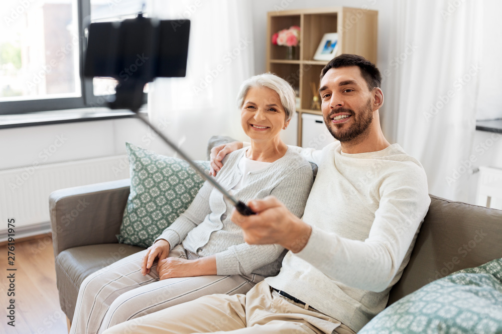Obraz premium family, generation and people concept - happy smiling senior mother with adult son taking picture by selfie stick at home