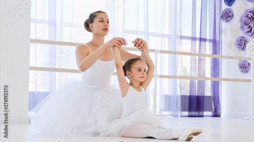 Little girl in white tutu and pointes is stretching on ballet lesson with teacher who helps small ballerina.