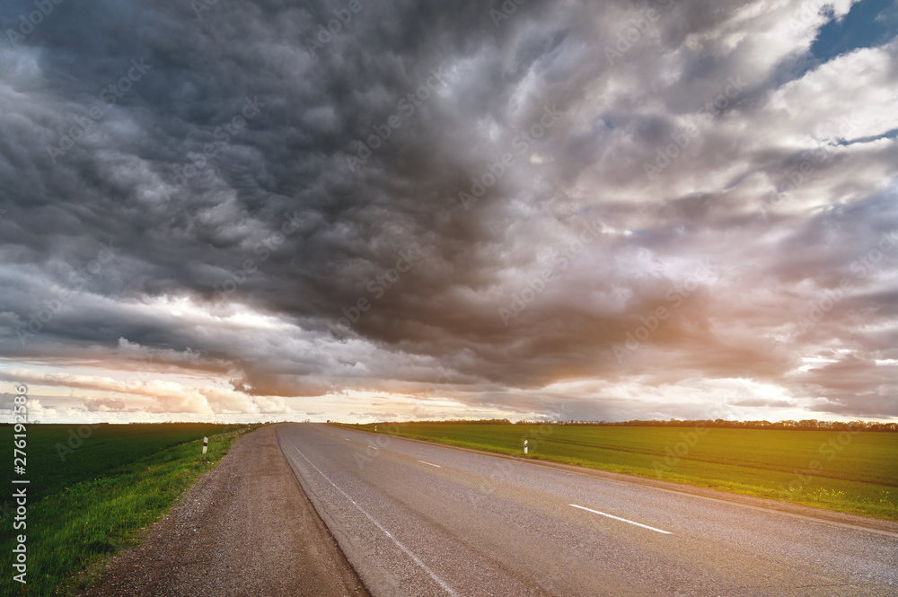Evening landscape of suburban asphalt road with low textured dramatic clouds and green fields on the sides