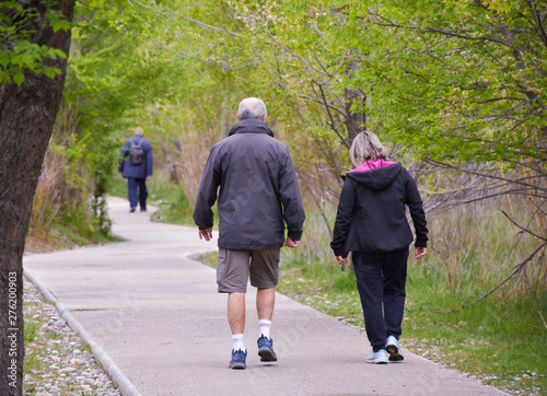 back view of a senior couple walking on a concrete way in the middle of a park. The male hiker wears sorts and a dark blue coat and the female hiker wears black tracksuit and coat. Horizontal picture