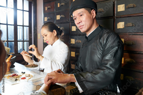 Traditional Asian medicine pratitioner checking pulse of customer when his coworker weighting treatment ingredients