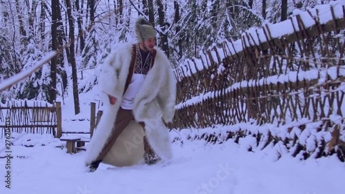 Slow motion of very cheerful or drunk young man in national wear and woolen coat walking in snow, sliding, entering the old wooden house. Customs and traditions, celebration