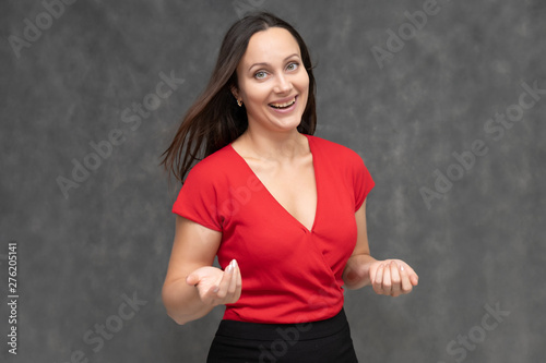 Portrait to the waist of a young pretty brunette woman of 30 years old in a bright red sweater with beautiful dark hair. It is standing on a gray background, talking, showing hands, with emotions