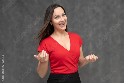 Portrait to the waist of a young pretty brunette woman of 30 years old in a bright red sweater with beautiful dark hair. It is standing on a gray background, talking, showing hands, with emotions