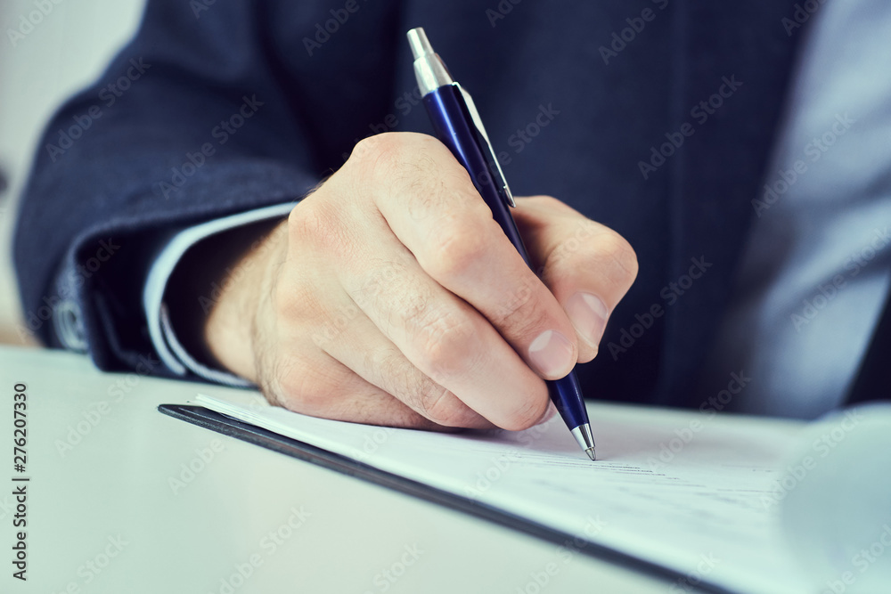 Hand of businessman in suit filling and signing with blue pen partnership agreement form clipped to pad close-up. Management training course, some important document, team leader ambition concept. Hand of businessman in suit filling and signing with blue pen partnership agreement form clipped to pad close-up. Management training course, some important document, team leader ambition concept.