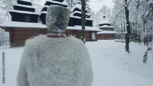 Rotation close up view of bearded young man in embroidered clothes and warm woollen coat standing in snow in front of the old wooden church breathing hardly. Religion, holiday