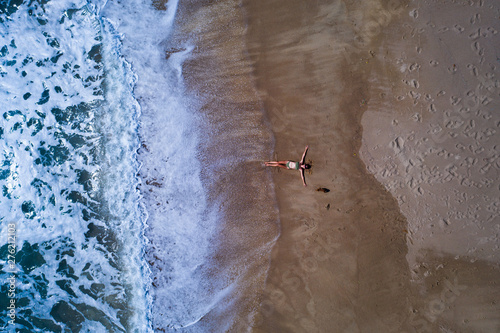 Aerial top view of woman with her hands outstretched laying on the sandy beach near the ocean. Summer holiday concept