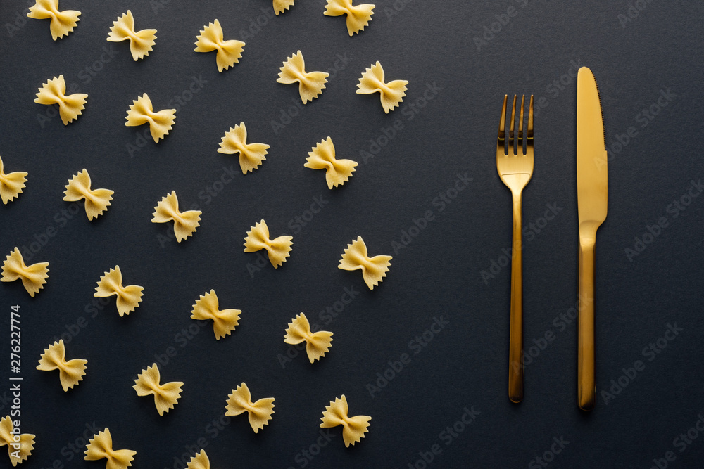 flat lay of farfalle pasta near golden knife and fork on black background - obrazy, fototapety, plakaty 