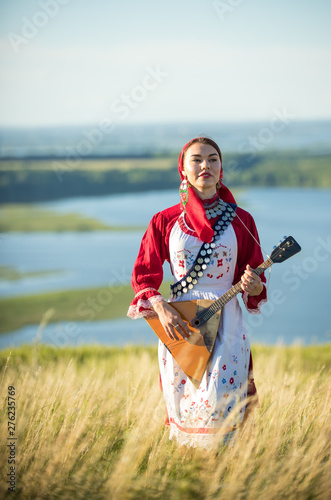 Fotografie A young woman in russian folk clothes standing on the field holding balalaika