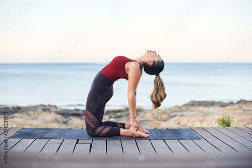 Young attractive woman practices yoga in camel pose on the beach.