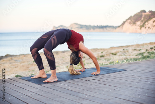 Young attractive woman practices yoga in wheel pose on the beach.