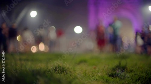 Green grass. A colourful crowd mowing at the background. People walking around. Creative evening lights and blurred shadows moving in the twilight. Festival in Barcelona.