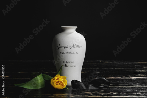 Fotografie Mortuary urn with ribbon and flower on table against dark background