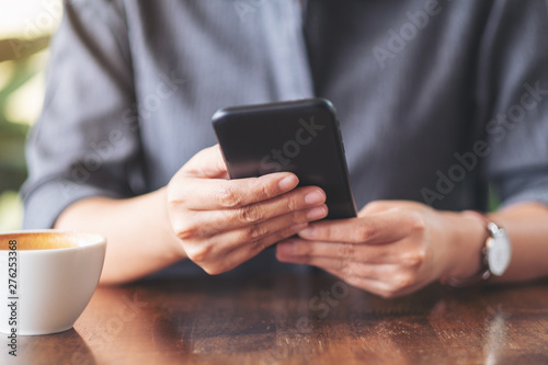 Closeup image of a woman holding  and using mobile phone with coffee cup on wooden table in cafe