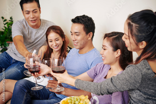 Group of cheerful friends sitting on sofa and toasting with wine glasses