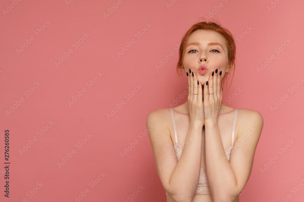 Young red-haired woman wearing lace bra gives an air kiss posing over pink background Young red-haired woman wearing lace bra gives an air kiss posing over pink background