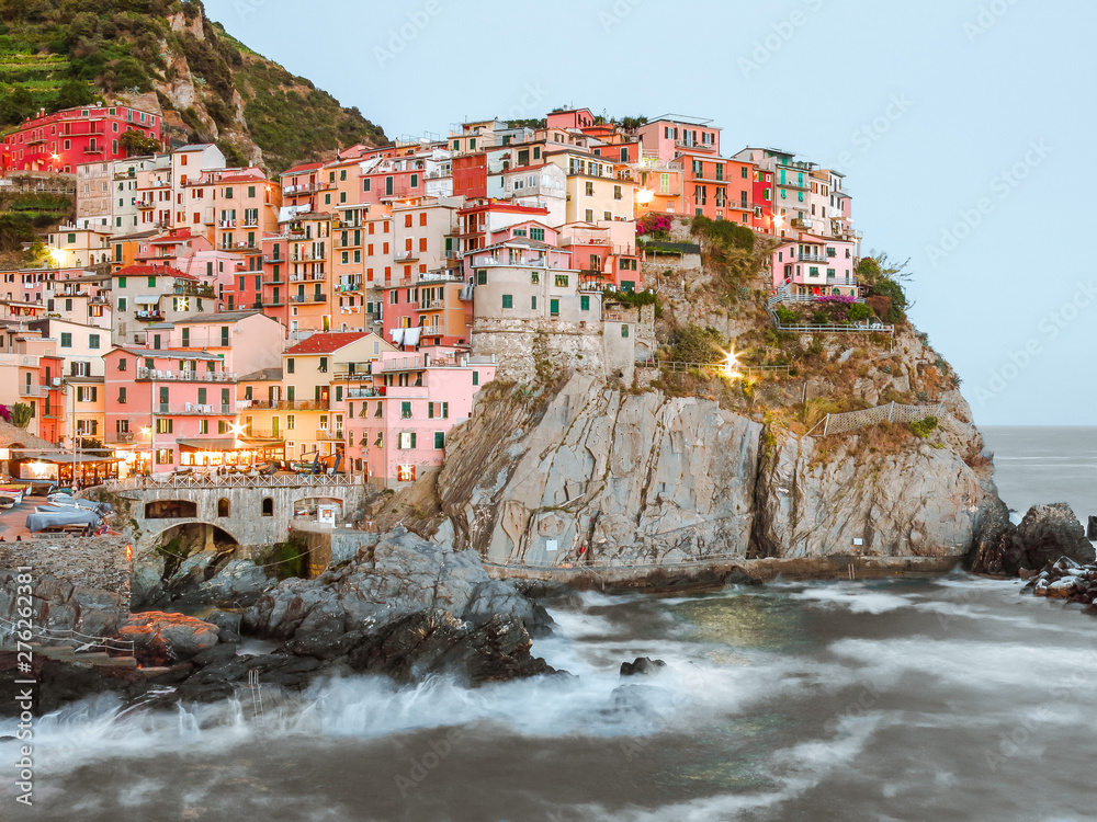 Colorful pastel houses along the cliffside in Manarola, Cinque Terre taken during the sunset with waves crashing along the coastline and a blue sky.