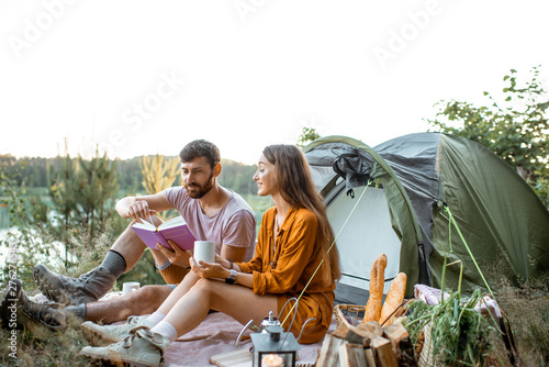 Young and cheerful couple sitting with book near the tent, enjoying the summer vacations in the forest during the sunset