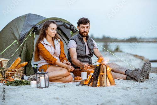 Young and cheerful couple sitting at the fireplace, cooking sausages, having a picnic at the campsite on the beach in the evening