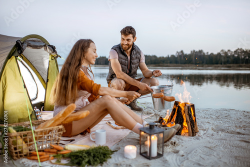 Young and cheerful couple cooking sausages at the fireplace, having a picnic at the campsite on the beach in the evening