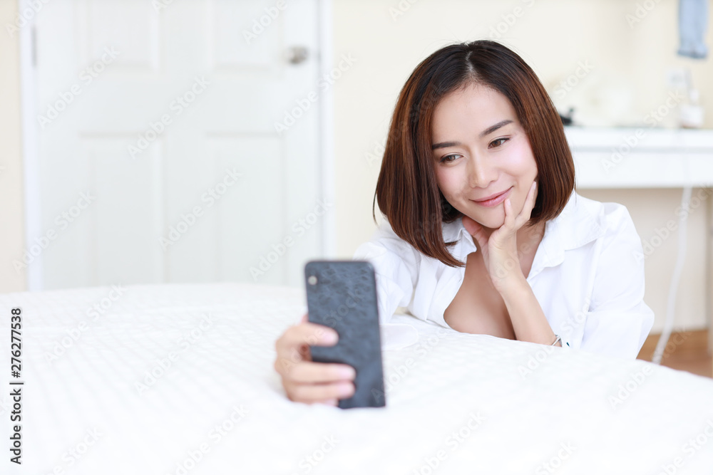 closeup beautiful asian woman, short hair in white shirt using video call or self portrait from mobile phone on white bed in bedroom with happy smiling face closeup beautiful asian woman, short hair in white shirt using video call or self portrait from mobile phone on white bed in bedroom with happy smiling face