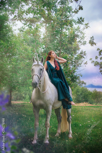 An elf with a unicorn. Beautiful girl in a green dress in nature with a white horse. Model in a medieval dress