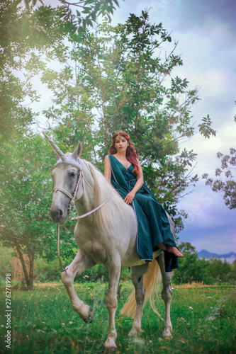An elf with a unicorn. Beautiful girl in a green dress in nature with a white horse. Model in a medieval dress