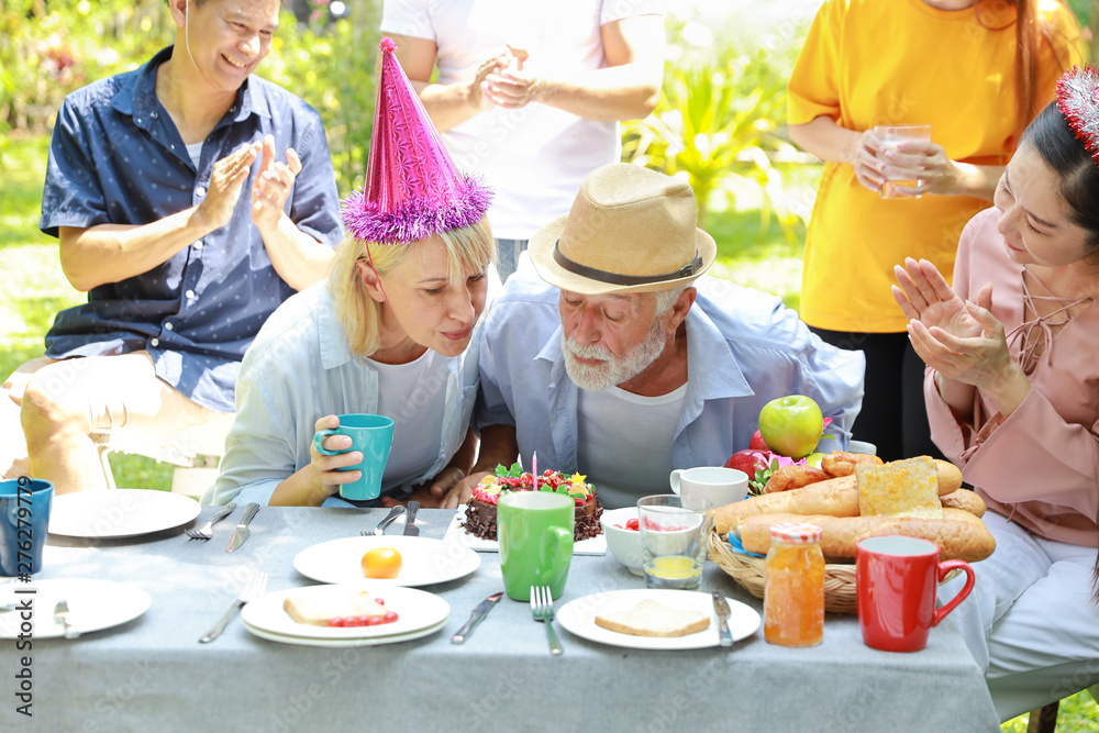 Happy multiethnic family giving surprise gift to caucasian grandfather on his happy birthday and he blowing out candles on homemade baked cake with happy face in backyard outdoor on sunny day
