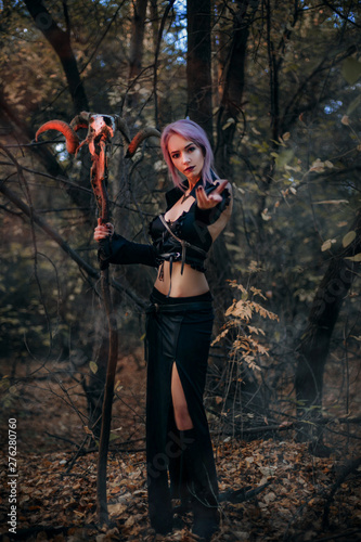 A witch with a staff from the skull of a ram conjures in the forest. A girl in a black dress and purple hair. Image for halloween