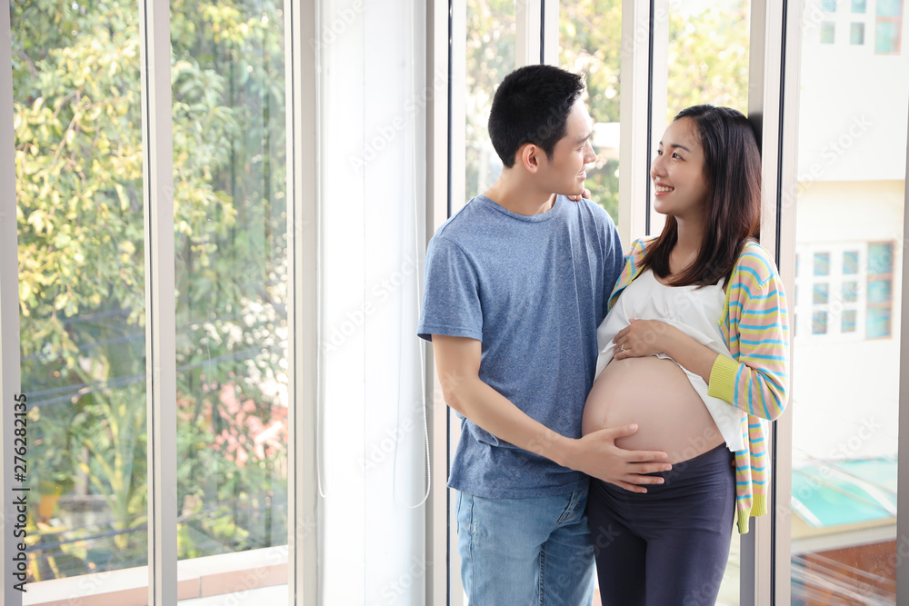 young asian husband standing and embracing, eyes contract together with young pregnant belly wife with happy smiling face and green trees background young asian husband standing and embracing, eyes contract together with young pregnant belly wife with happy smiling face and green trees background