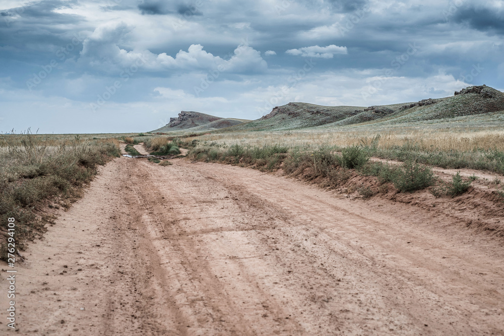 The clay road in the steppe among the hills against the backdrop of a dramatic leaden sky.