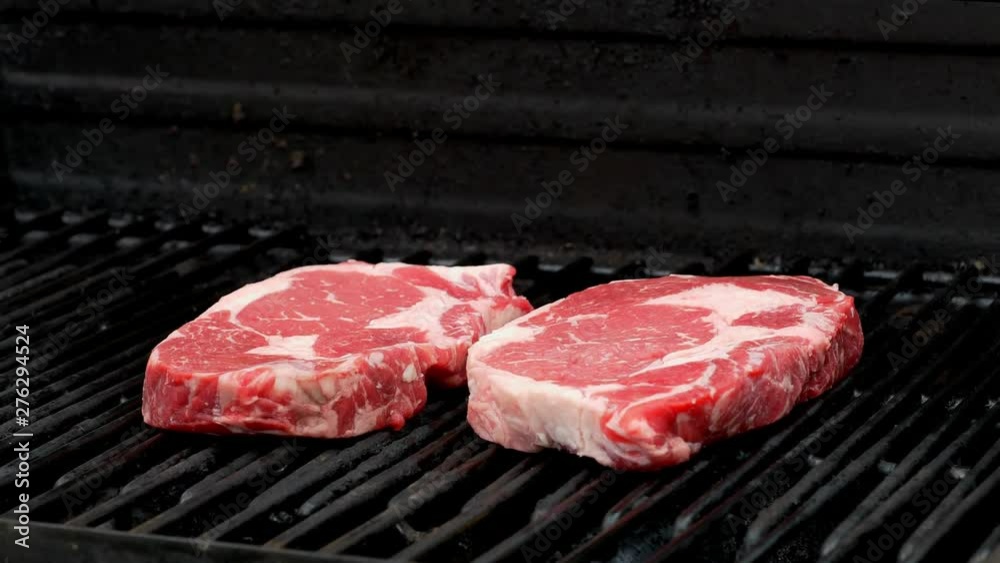 A raw rib eye steak is placed on a grill next to another one. A raw rib eye steak is placed on a grill next to another one.