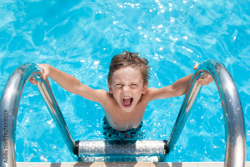 happy delightful small kid with wet hair standing on chrome handrail in fresh water swimming turquoise outdoors pool in sunny summer holiday leisure day with copy space