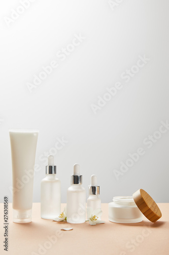 cream tube with cream  cosmetic glass bottles with serum  open jar with wooden cap and few jasmine flowers on beige