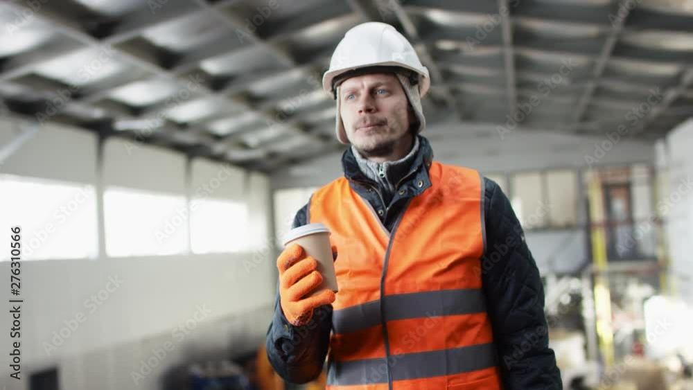 man beard and mustache in a hard hat and overalls is standing in the middle of the hangar and drink coffee at the camera